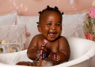 Smiling baby sitting in a bathtub, playing with bubbles and soap on skin.