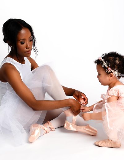 Woman in a white ballet dress helping a young girl in a pink tutu tie a ribbon.